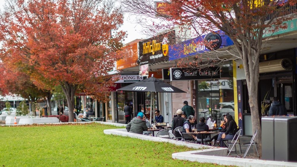 Photo of people sitting out outdoor tables in front of business along Hampshire Road in Sunshine. There is gree grass in the foregroundand trees with red leaves to the left.