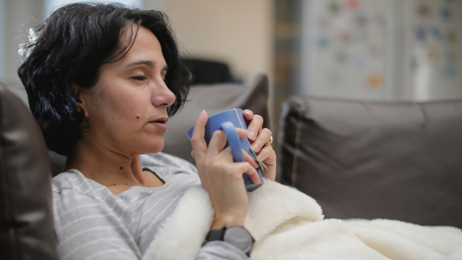Woman lying down on couch covered with a blanket holding a hot drink