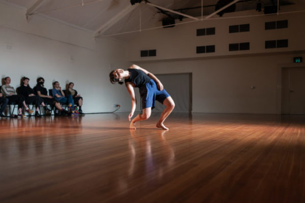 A dancer performs in a wooden floored space, severalaudience members are seated at one side of the room