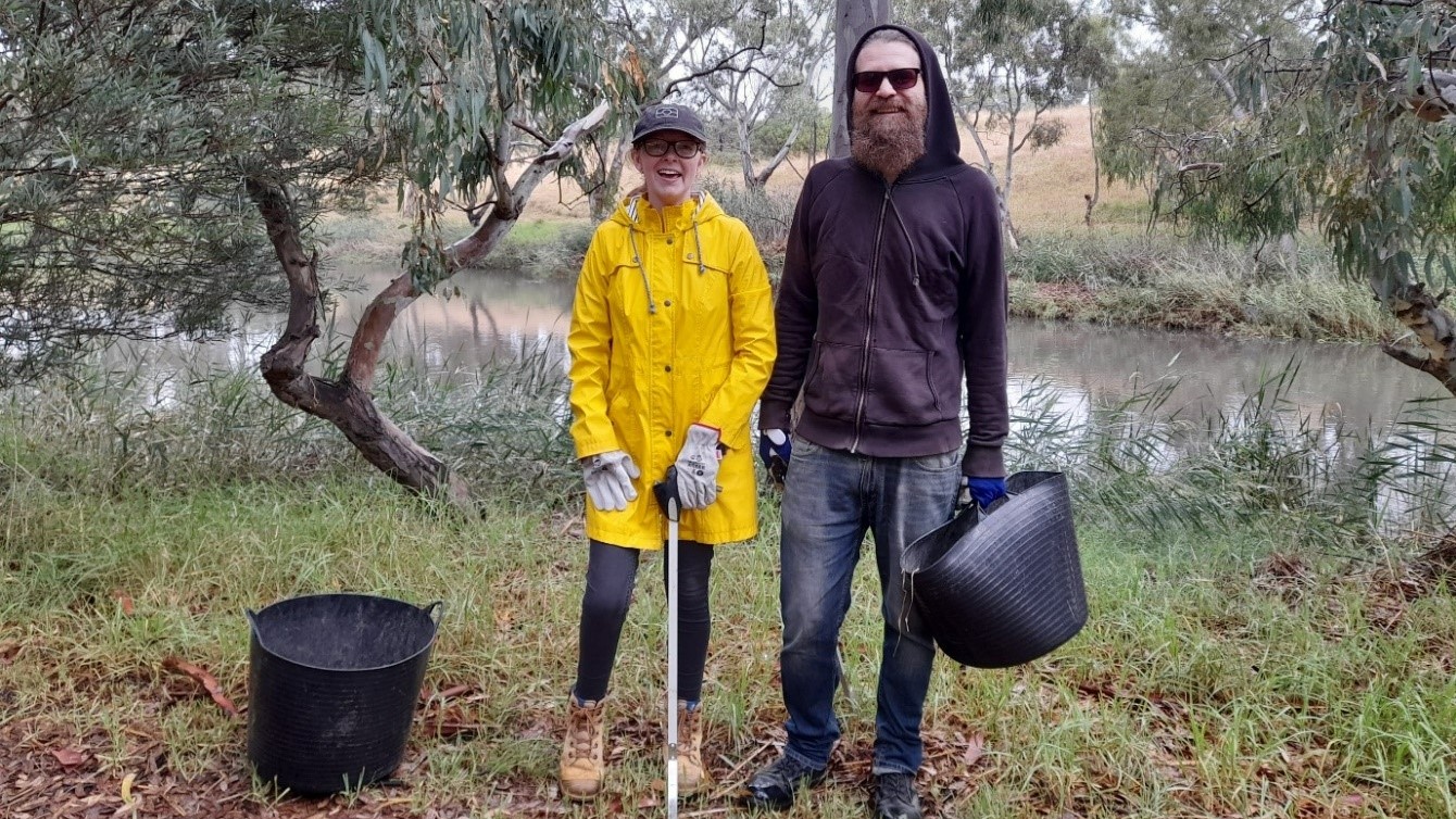 Two volunteers stand in front of a creek. The woman on the left wears a cap and yellow raincoat. A man on the right wears a dark coloured hoodie and sunglasses and hold a plastic bucket in his left hand.