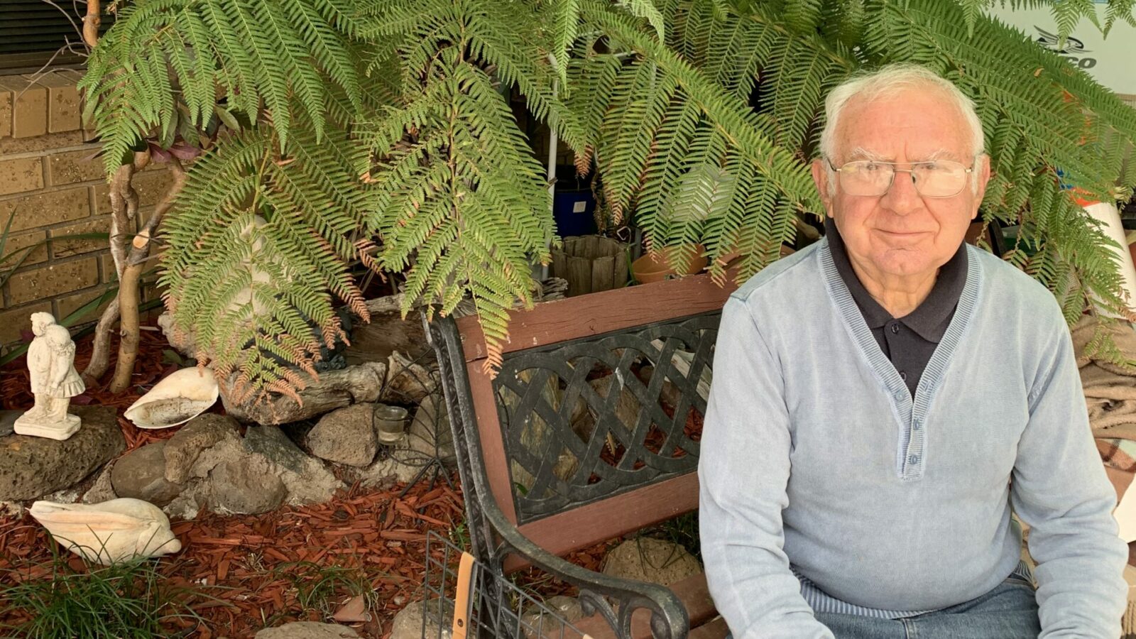 Elderly gentleman sitting on a bench in front of ferns and garden ornaments.