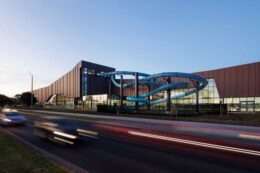 Picture shows the Brimbank Aquatic and Wellness Centre at dawn or dusk with a clear view of the slide coming out of the wall of the building and cars in the foreground travelling at high speed and blurry.