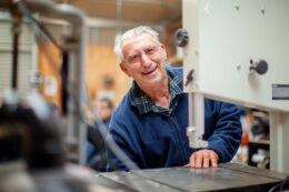 A man with grey hair smiles at the camera from behind some machinery in a workshop or learning space.
